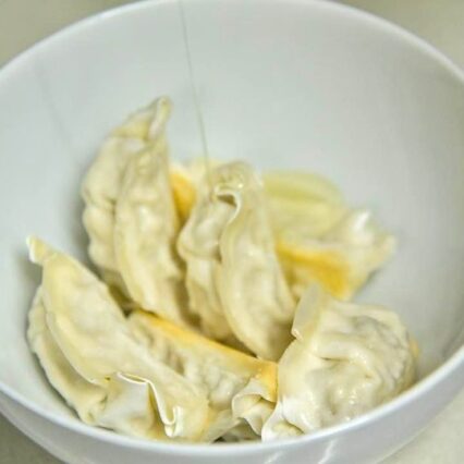 Frozen gyoza in a white bowl being drizzled with oil
